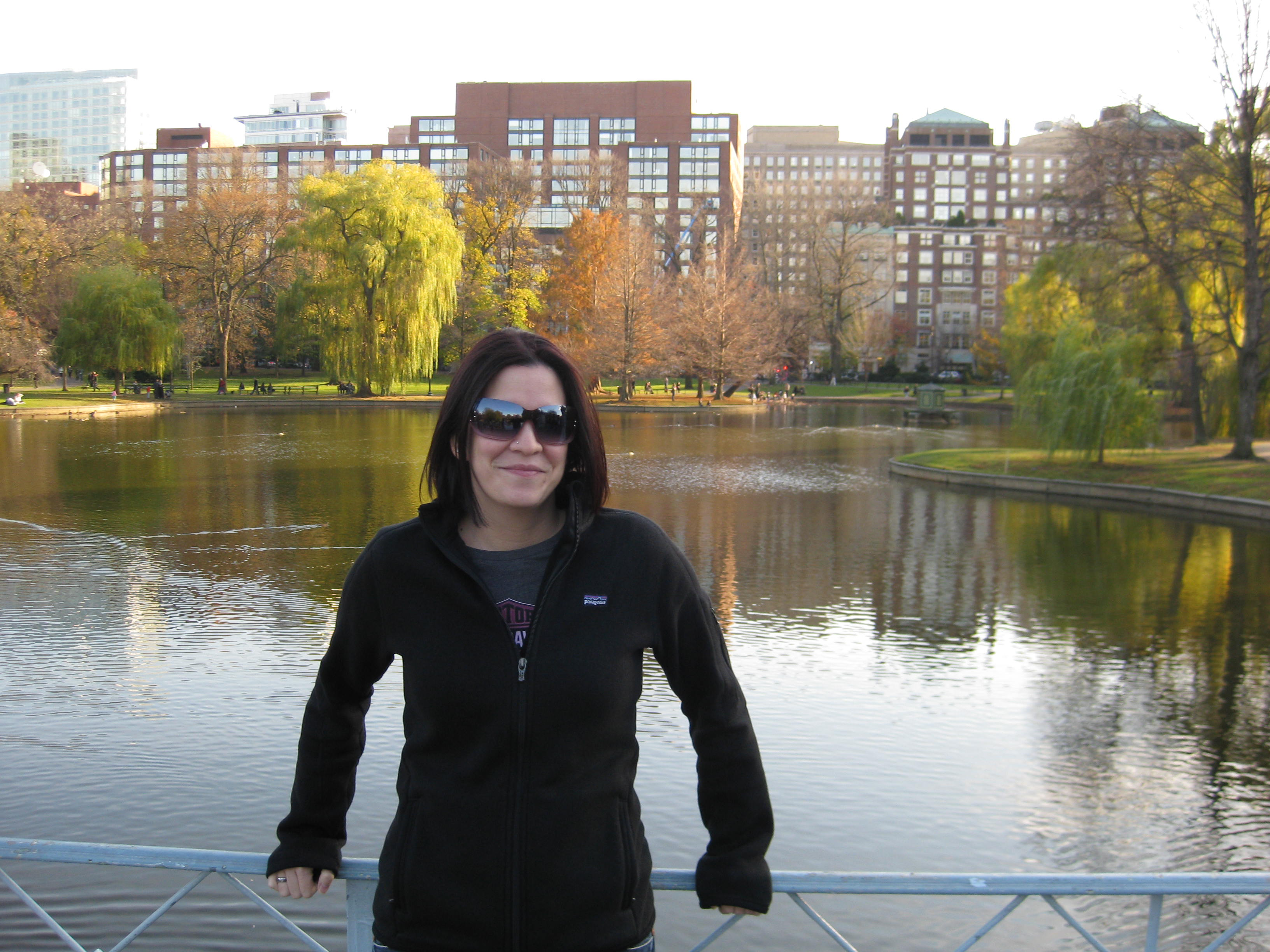 A brunette woman in a black zippered sweater wearing sunglasses and standing in front of a body of water with trees in the background.
