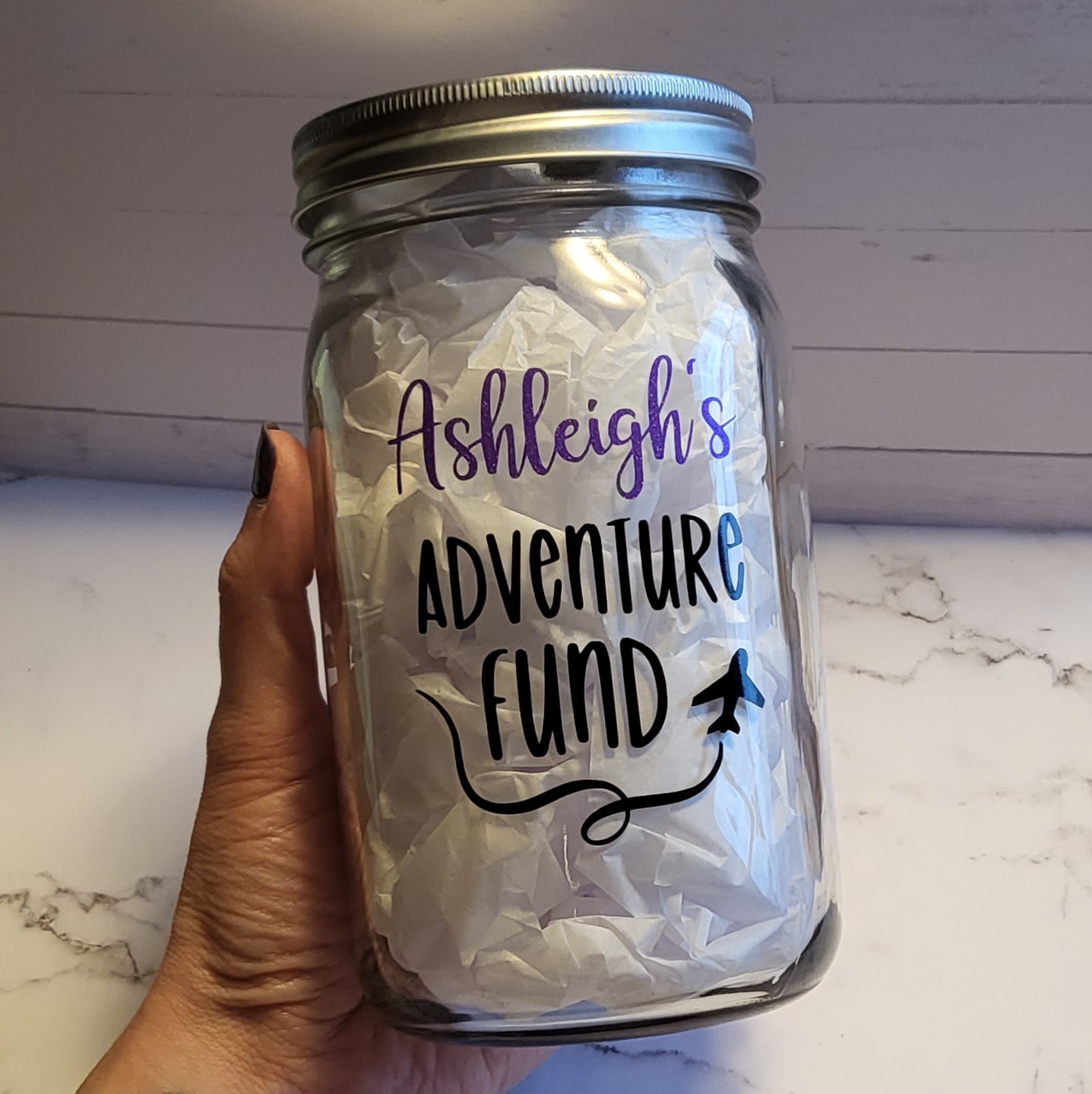 A 32 oz mason jar that has custom-made design in vinyl lettering that says Ashleigh's Adventure Fund.