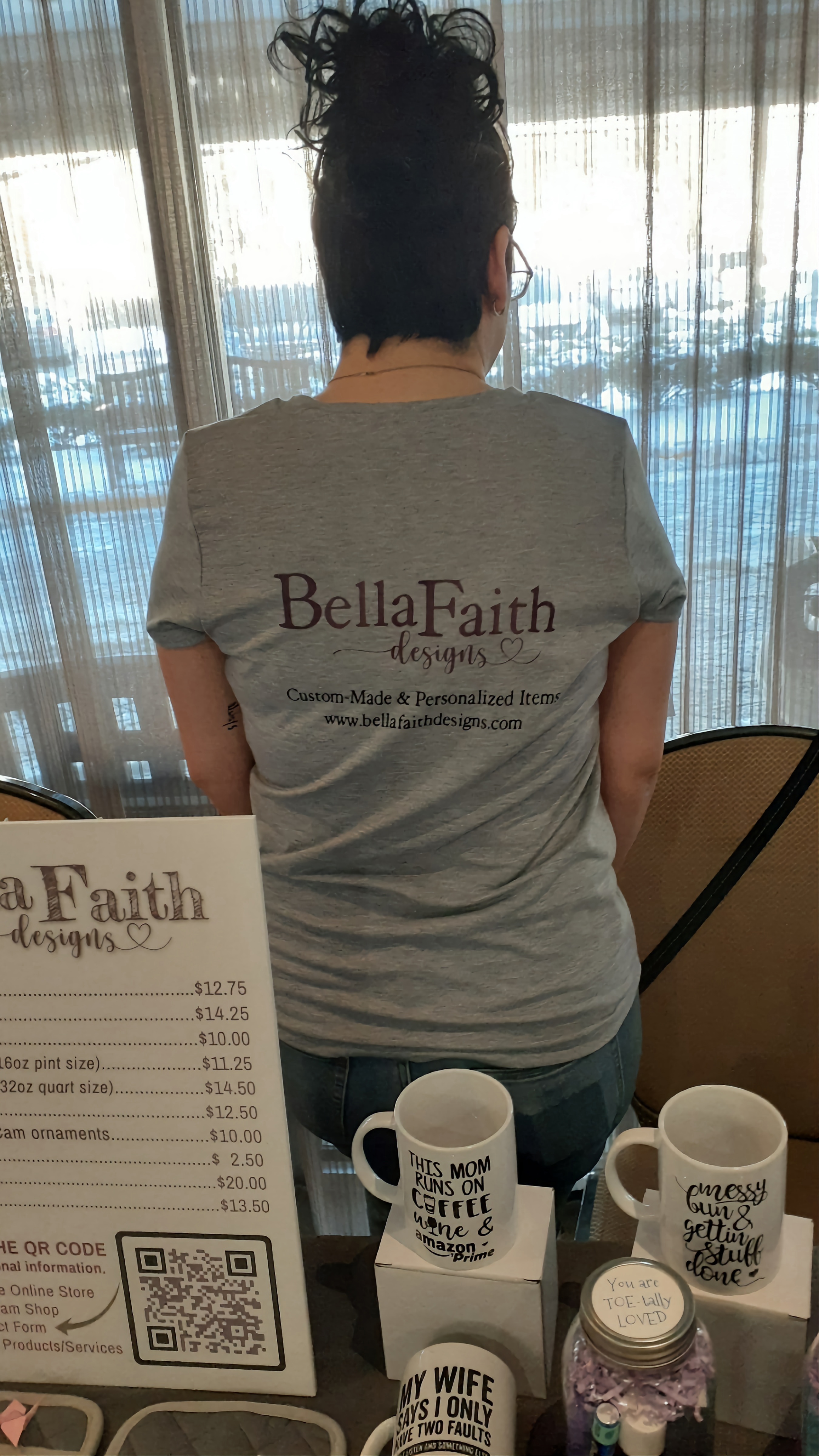 A brunette woman facing the other way at a event showcasing her products wearing a shirt that says Bella Faith Designs on the back.
