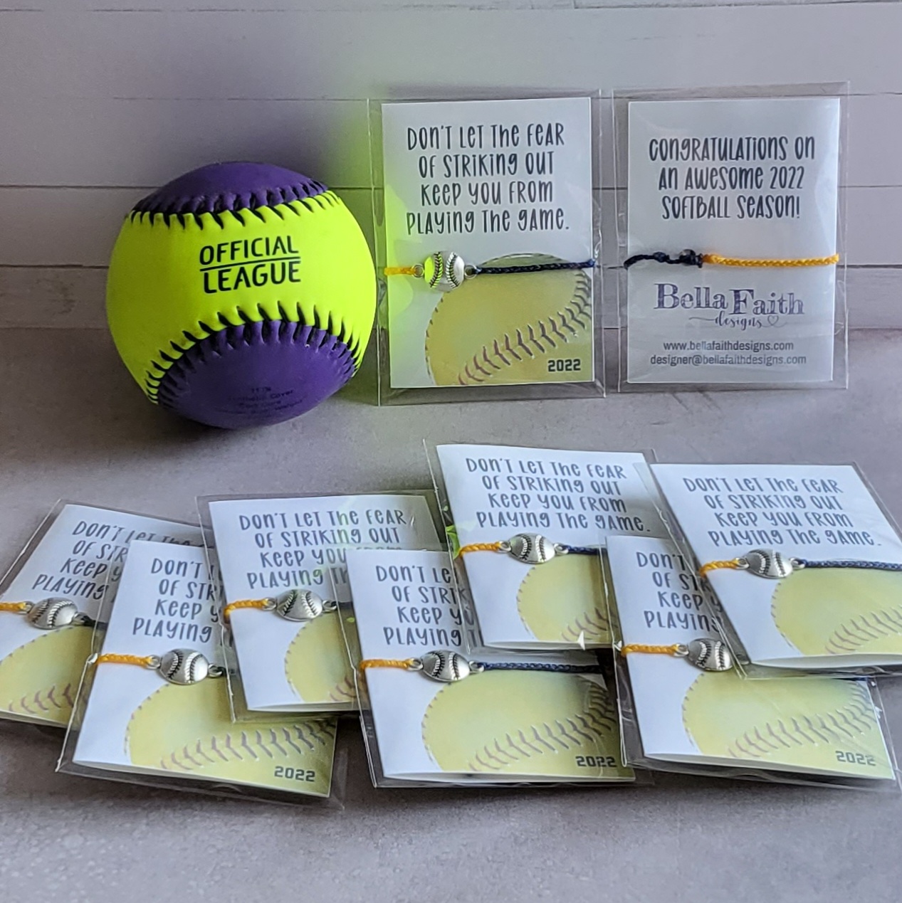A yellow and purple softball next to favors that have two-toned yellow and blue corded bracelets with a softball charm.